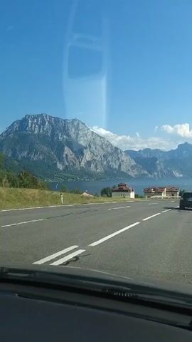 This is a first-person perspective video captured from inside a vehicle driving on a road. The road is paved with lane markings and appears to be a multi-lane highway. On the left side of the road, there is a body of water, likely a lake, bordered by a steep, mountainous landscape with a clear blue sky overhead. Several buildings with red roofs are visible along the lakeshore. Cars are driving in the same direction as the video taker, and some cars are visible in the distance on the opposite side of the road. The overall scene suggests a scenic drive through a mountainous region with a lake.