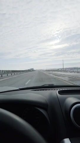 The video shows a point-of-view shot from inside a vehicle driving on a highway. The sky is overcast with textured clouds. The road ahead appears wet, and the roadside is lightly covered in frost or snow. Guardrails line the left side of the road, and fields stretch out on either side. A few other vehicles can be seen in the distance on the highway. The vehicle's dashboard is visible, including the steering wheel and parts of the instrument panel. The general environment suggests a cold, possibly winter, day.