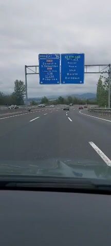 A dashcam view shows a car driving on a multi-lane highway under a cloudy sky. A large blue overhead highway sign displays directions to various cities including Donostia, S. Sebastian, Lasarte-Oria, Iruna, Pamplona, Hernani, Passai, Irun, and Bordeaux. The car is in the middle lane, with other vehicles visible ahead and on adjacent lanes. The environment appears to be rural, with trees and hills visible in the distance. The road is asphalt, with white lane markings. The weather is overcast.