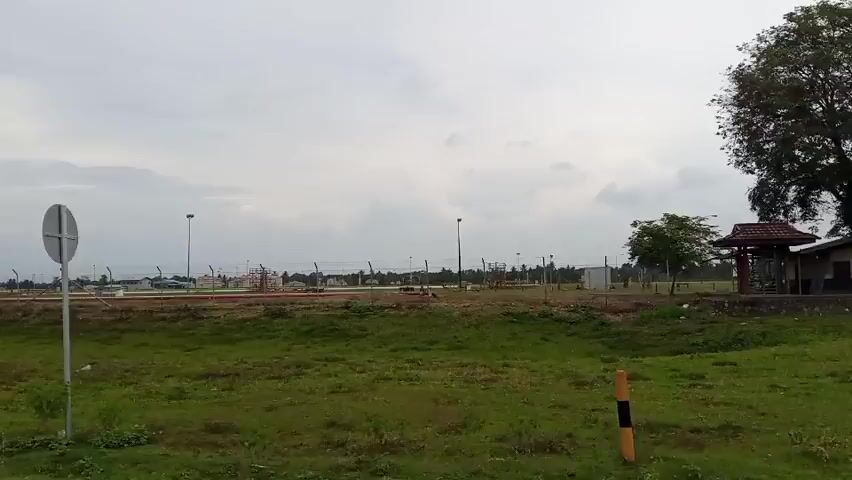 The video pans across a landscape under an overcast sky. In the foreground, a patch of green grass and dirt is visible. A white signpost stands in the middle ground. Behind the signpost, a long stretch of barbed-wire fencing runs horizontally across the frame. Various structures, including what appear to be industrial buildings or storage units, are visible behind the fence. Several tall light poles are interspersed along the fence line. To the right, a small building with a brown roof is partially visible. The background consists of a distant treeline and the cloudy sky.