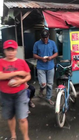 The video shows an outdoor scene, likely a street or market. In the foreground, a person wearing a red t-shirt and a red baseball cap is holding a small object, possibly food, and looking at the camera. Their face is slightly blurred due to motion. To their left, another person's legs are visible, seated or squatting. In the background, a man wearing a blue polo shirt, jeans with ripped knees, and a black baseball cap is standing near a food stall. He is holding what appears to be money in his hands. A bicycle, light blue in color, is parked in front of the food stall. The stall itself has a red awning and displays some signage. The ground is wet, suggesting it may have recently rained. The overall impression is of a casual, everyday setting.