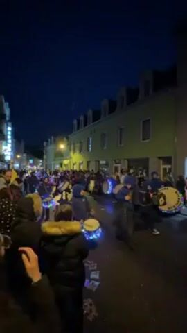 The video shows a crowd of people gathered on a street at night. In the middle of the street is a group of people performing, playing drums that are decorated with colorful lights. A few people are also playing accordions. The performers are all dressed in blue jackets and hats. The crowd of onlookers is dense, many wearing winter clothing. The street is lined with buildings, with one building distinctly painted a light green. The streetlights cast a warm glow on the scene.