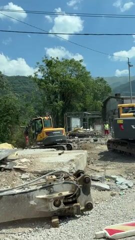 A construction scene with two excavators and a hydraulic breaker in the foreground. One excavator, a yellow Volvo, is to the left, and another, a dark gray Volvo, is to the right. Both are on a dirt and debris-covered surface. A person in a yellow vest and red pants stands between the two excavators, appearing to direct operations. In the background, trees and hills are visible under a blue sky with scattered clouds. The excavators are positioned near the remnants of a demolished structure, with debris scattered around. The hydraulic breaker, a large piece of equipment, is in the foreground, suggesting demolition or breaking up concrete.