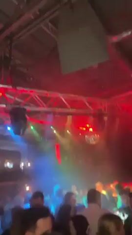 The video shows a densely packed dance floor in a nightclub. Red and blue stage lights illuminate the crowd. Above, a metal framework supports lighting equipment, and a disco ball hangs from the ceiling. People are standing close together, talking and facing different directions. The overall atmosphere is dark and energetic.
