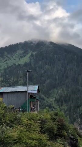 A wooden cabin with a metal roof sits on a hillside. Dense green trees cover the steep mountain slopes. Clouds obscure the peak of the mountain. A waterfall cascades down the mountainside in the distance.