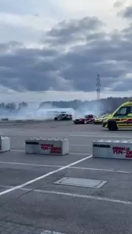 The video shows a drifting competition in an open parking lot on an overcast day. Several cars are performing drifting maneuvers, creating a large cloud of smoke. The cars are of various colors, including black, burgundy, and lime green. Concrete barriers are visible in the foreground, each labeled with "FUN-DRIVE" in red lettering. In the background, beyond the drifting area, there's a white tent-like structure and a lattice tower. An ambulance is parked to the side of the drifting area. A white flag is attached to a pole next to one of the barriers. The parking lot has white lines marking spaces.