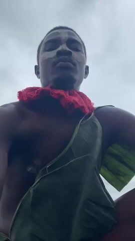 The media file features a young Black man outdoors, with a dramatic, overcast sky and a lush, green landscape suggesting a hilly or mountainous region. The man is adorned with white face paint, applied in a striking, symmetrical pattern across his forehead and cheeks, and a bright red, ruffled collar. He is also draped in a large green leaf, possibly a banana leaf, that functions as a garment. His skin is dark and his physique appears muscular.

The media captures the man in a series of close-up shots, highlighting his facial features and adornments. In one shot, he looks directly at the camera with a serious, perhaps stoic expression. In others, he turns his head, revealing the profile of his face and the extent of the face paint. The background shifts from a close-up of the sky to a wider view of the landscape, showcasing rolling green hills and vegetation. The overall impression is one of a person participating in a traditional or ceremonial activity, possibly a ritual or performance, set against a natural, untamed environment. The time of day appears to be overcast, likely afternoon, given the diffused light. There are no visible text elements in the media.