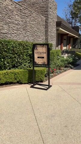 The video opens with a sign for EREWHON THOUSAND OAKS NOW OPEN 7AM - 9PM DAILY. The sign is on a black stand in front of a building with a stone facade and a landscaped area with bushes and rose bushes. The camera pans to the right, showing a pathway and a person walking away from the camera. The pathway is made of light-colored aggregate and bordered by a darker aggregate. The building continues to the right, with a brown awning and decorative lamps. The person walking away is wearing dark clothing and a backpack. The scene is outdoors during the day, with clear blue skies.