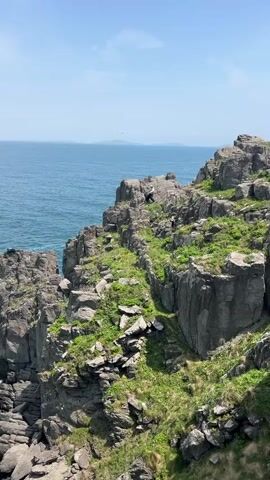 A medium, high-angle shot shows a rocky, grass-covered seaside cliff on a bright, sunny day. Two people, appearing small in the frame, navigate the rocks on the cliff. The sea extends to the horizon, with a few small islands visible in the distance. The sky is mostly clear, with a few scattered clouds. Waves crash against the base of the cliff.