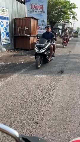 The video shows a person riding a black scooter on a paved road. They are wearing a blue cap and a dark t-shirt. The scooter has gold rims. Another person is riding a red motorbike in the background. There are buildings and signs on the side of the road, including one that says "Wakaf Masjid Al Hidayah." The road appears to be in a somewhat urban or semi-urban area with some signs of commercial activity. The lighting suggests it is daytime.