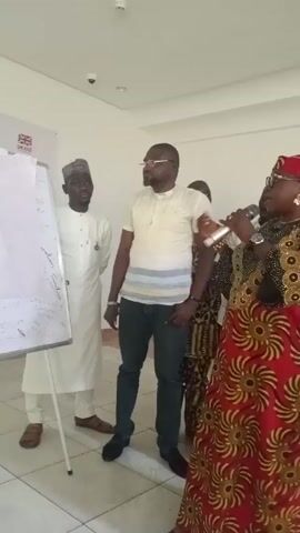 The video shows a woman speaking into a microphone. She is wearing a red and gold patterned dress with a matching head wrap. There are several other people in the room, including two men standing next to a whiteboard. One of the men is wearing a white robe and a hat. The other man is wearing a white shirt with a striped collar and jeans. The room is brightly lit and has white walls. The woman speaking is gesturing with her hands as she talks. The other people in the room appear to be listening attentively. She is discussing extortion and bribery punishment.