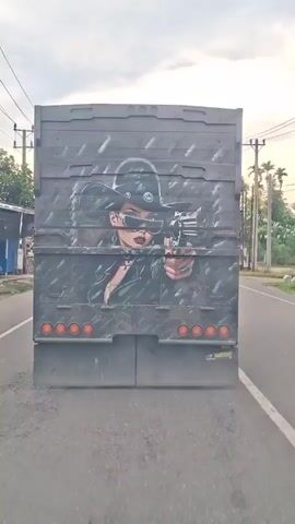 A truck travels down a road, its rear door featuring an illustration of a woman wearing a cowboy hat and sunglasses, aiming a gun. The woman is depicted in a leather jacket with visible cleavage and jewelry. The truck is gray with a textured background behind the illustration and features orange reflector lights. The road is asphalt and has white lines on either side. Palm trees and buildings are visible in the background, with utility poles lining the road. The sky is overcast with a hint of sunlight breaking through. The "WARRIORS" label is visible on the lower right corner of the truck's rear door.