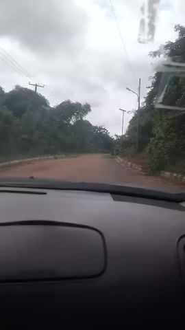 This media file shows a point-of-view perspective from inside a vehicle traveling on a dirt road. The scene is rural, with lush green trees and vegetation lining both sides of the road. In the distance, a power line pylon is visible, indicating the presence of electricity infrastructure. The road itself appears to be unpaved and somewhat uneven.

The weather is overcast, with a cloudy sky suggesting it might be humid or that rain is possible. The time of day is indeterminate due to the cloud cover, but it appears to be daylight. The overall atmosphere is quiet and somewhat isolated, typical of a rural setting.

Notable details include a street lamp on the right side of the road and what appear to be stacked concrete blocks or building materials near the roadside. The interior of the car is visible in the foreground, specifically the dashboard, with "SRS AIRBAG" text visible on it. There are no people visible in the frame, only the road and the surrounding natural and man-made elements. The activity is simply driving through this environment.