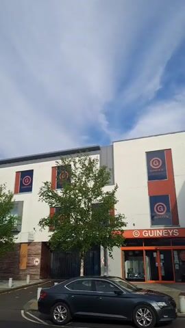 A video recording pans across the exterior of a modern, multi-story building with a white facade. Several rectangular banners with a circular logo in red and blue are visible on the upper level. Trees with green foliage are planted in front of the building. The ground level features large windows and a sidewalk. Cars are parked in front of the building and on the street. The sky is partially cloudy.