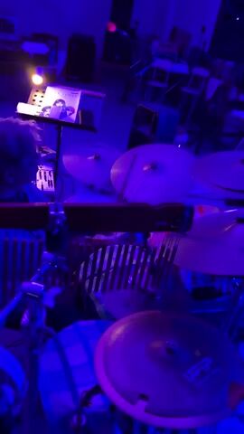 A person plays drums, while another person plays the keyboard, in a room lit by purple lights. The drummer is mostly obscured by the drums, but their hands are visible, holding drumsticks and hitting the drums. The keyboard player is visible in the background, sitting at a keyboard and playing it. The room is dimly lit, with the purple lights casting a glow over the instruments and the musicians. There are also other instruments and equipment visible in the background, including a microphone and a music stand.