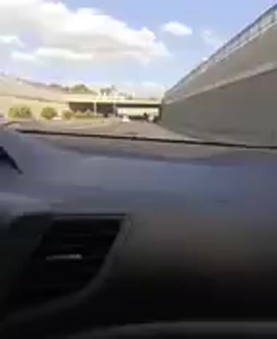 The media file provides a first-person perspective from inside a vehicle. The view is directed forward through the windshield, revealing the dashboard of the car prominently in the foreground. The dashboard is dark gray and includes an air vent on the left side. Beyond the car's interior, the scene unfolds outdoors.

The setting appears to be an urban or suburban environment, possibly near a highway or overpass. A large, elevated concrete structure, likely part of a bridge or overpass, dominates the middle ground. It spans across the view, casting shadows on the road below. In the distance, more structures and buildings are visible, suggesting a developed area. The sky above is partly cloudy, with patches of blue sky visible between the white clouds, indicating it is daytime, likely late morning or afternoon given the brightness. The lighting suggests fair weather.

There are no people clearly visible in the media file. The primary objects in focus are the car's interior, the overpass structure, and distant buildings. The activity taking place is driving, as indicated by the perspective from within a moving vehicle. The overall mood is neutral, focusing on the visual elements of the drive. No text is visible in the media file. The location context provided is Mamak, Turkey.
