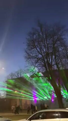 At dusk, green and purple lasers illuminate a building while a crowd of people watch.  Streetlights glow in the foreground and background.  Bare trees line the street. A white car is parked in the foreground.  The camera moves upward, eventually focusing on a light pole.