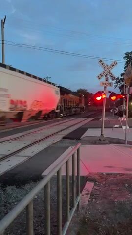 A train crosses railroad tracks at a crossing. A railroad crossing signal with flashing red lights is visible. The train is an orange and black locomotive pulling a white car. The sky is blue with some clouds. Trees are visible in the background.