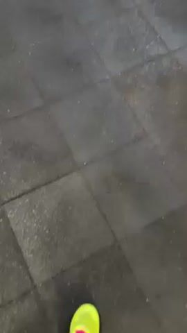 The video shows a person's foot wearing a bright green and purple sneaker on a dark gray, textured floor with square tiles. The camera is positioned looking down at the foot and the floor. A hand with a gold bracelet appears, making the "okay" sign. The hand has a visible scratch on the skin near the wrist. The background shows a glimpse of a purple wall and a dark, possibly black, object to the right.
