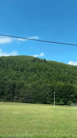 The video shows a wide shot of a grassy field with a tree-covered hill in the background under a clear blue sky with a few wispy clouds. A single utility pole stands in the field. The camera appears to be moving, possibly from a vehicle, as indicated by the slight motion blur and the way the utility pole moves in relation to the background. There are no people visible in the video.
