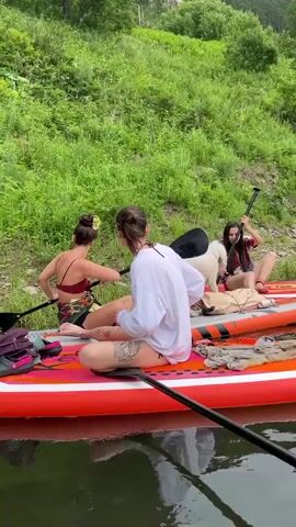 Three young women and a white poodle are on paddleboards in the water. The woman in the foreground is wearing a white oversized shirt and shorts. The woman behind her is wearing a bikini and shorts. The woman on the right is wearing a patterned top and shorts. The poodle is standing on the middle paddleboard. The women are holding paddles. They are near a grassy bank with trees. The water is dark and reflective. The paddleboard in the foreground has a fish design.