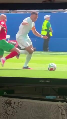 A soccer player in a white jersey with the number 10 and orange accents on his sleeve is dribbling the ball. A player in a red and green uniform lunges to tackle him. The player in white attempts to evade the tackle by stepping over the ball. As he does, the tackling player's left leg makes contact with the player in white's right leg, causing the player in white to fall to the ground. The ball rolls away from both players. In the background, a security guard in a yellow vest is visible, along with spectators in the stands. The scene takes place on a bright, sunny day on a green soccer field.