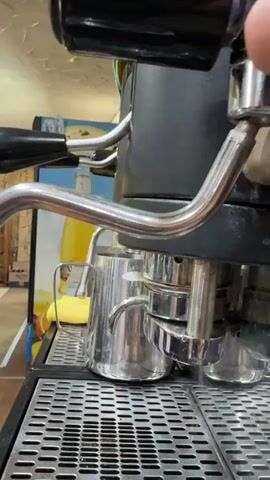 A close-up, side view shows an espresso machine. A metal pitcher sits on the grates of the machine below a chrome spigot. A hand is visible at the top right of the frame, operating a lever on the machine. Dark liquid begins to flow from the spigot into the pitcher.