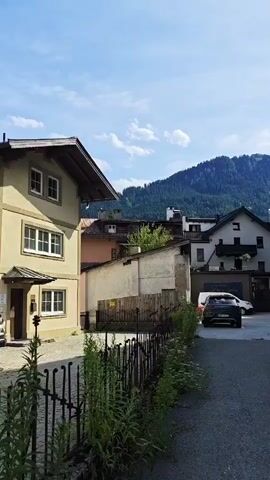 This is a vertical, medium shot of a street in a village or town, with buildings on either side and a mountain in the background. The sky is blue with some clouds. The buildings appear to be a mix of residential and commercial, with some having balconies and decorative trim. There are several cars parked on the street and in a driveway. A black metal fence runs along the left side of the street, with some greenery growing through it. The road is paved with asphalt. The video is still and there is no visible movement or action.