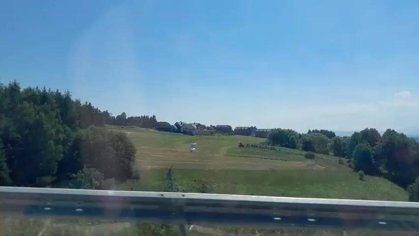 The video shows a view from a moving vehicle, looking out at a rural landscape on a sunny day. In the foreground, a metal guardrail is visible. Beyond the guardrail, there are rolling green fields, some of which appear to be recently harvested, with scattered bales of hay. A line of trees runs along the left side of the frame, and scattered trees are also present in the fields. In the distance, a cluster of houses with dark roofs is situated on a hill. The sky is clear and blue, with a few wispy clouds. The overall impression is one of a peaceful, pastoral setting.