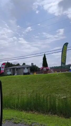 The video is shot from inside a vehicle, looking out the side window and the rearview mirror. The exterior scene features a grassy embankment with tall green grass in the foreground. Several Canadian flags on poles are visible, some with a red maple leaf design. Buildings, including houses and what appears to be a commercial establishment, are situated on the other side of the embankment. Power lines and poles crisscross the sky, which is cloudy. A person's arm and hand, holding a phone, are reflected in the rearview mirror. The vehicle appears to be moving, indicated by the slight blur of the background. There are no other people or distinct actions visible in the video. The overall environment suggests a rural or suburban setting in Canada, possibly during the daytime.