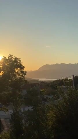 The video shows a scenic landscape at sunset. In the foreground, trees with dark green foliage frame the view. The sun, a bright orb, is setting on the left side of the frame, casting a warm golden glow across the sky and illuminating the leaves of a large tree. Beyond the trees, a small village with red-roofed houses is nestled in a valley. In the distance, rolling hills and mountains recede into the hazy horizon, painted in soft shades of purple and blue. A body of water, possibly a lake or bay, is visible in the mid-ground, reflecting the warm light of the sunset. Power lines with insulators are visible on the right side of the frame, stretching across the scene. The sky is a clear gradient, transitioning from a pale blue at the top to a vibrant orange and yellow near the horizon. The overall atmosphere is peaceful and serene, capturing the beauty of a natural landscape at dusk.