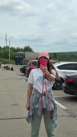 The video begins with a young woman in a pink cap and white t-shirt taking a selfie with her pink smartphone. She is wearing a plaid shirt tied around her waist and green pants. She is standing in a parking lot with cars and a large truck in the background. The scene then transitions to a wider view of the parking lot and the road beyond. A few people are walking in the distance, and a child in a blue shirt and green pants is seen walking up a grassy incline towards a large sign. The sign appears to be a landmark or entrance marker, featuring the Russian flag colors. The video pans to show more of the sign and the surrounding green landscape.