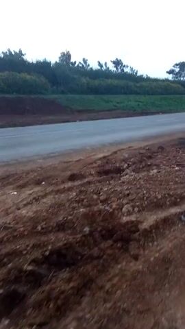 The video shows a car driving on a paved road. The camera is positioned on a dirt slope beside the road. The car drives past the camera, then away into the distance. The surrounding environment includes green fields and trees. The sky is overcast.
