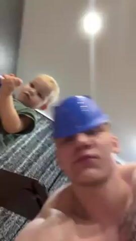 A shirtless man with a blue plastic hat and a tattoo on his chest is shown in an indoor setting. A child is also present, holding a blue toy gun with white foam on it. The child is looking directly at the camera, while the man seems to be focusing on something else. There are curtains in the background. The man asks "Что чиним?" (What are we fixing?) and then "Работа идёт?" (Is the work going?) suggesting they might be engaged in a repair activity together.