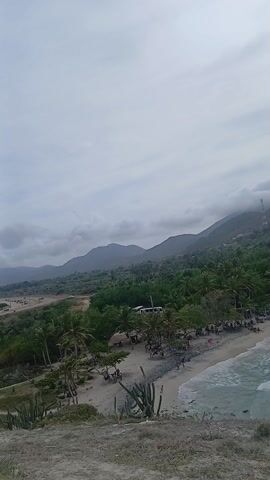 The video shows a high-angle view of a crowded beach on a cloudy day. The beach is lined with palm trees and buildings, and people can be seen swimming, sunbathing, and walking along the shore. The waves are small and the water is a greenish-blue color. In the background are mountains covered in green vegetation. The overall scene appears to be a popular tourist destination.