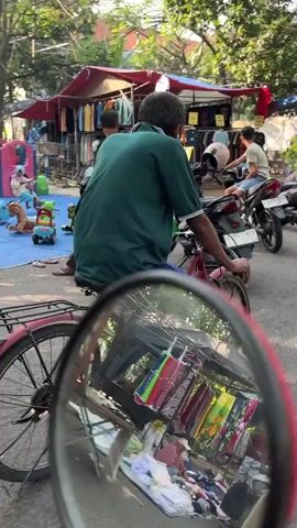 The video opens with a reflection in a red bicycle mirror, showing clothes hanging on a line and piles of fabric on the ground. The camera then pans to the right, revealing a busy street scene. A man in a green shirt is seated on a bicycle, facing away from the camera. To his right, another man is sitting on a motorcycle, and further to the right, there are more motorcycles parked. In the background, there are market stalls with red and blue awnings. One stall displays various colorful clothes. There are also children's toys visible, including a rocking horse and a small blue and yellow ride-on toy. The environment appears to be an outdoor market, possibly in a developing country, with a mix of everyday life and commerce. The lighting suggests it is daytime, with sunlight filtering through trees.
