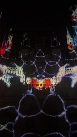 A light show is projected onto a building at night. The lights create abstract patterns and shapes in various colors, including red, white, and blue. The patterns shift and change throughout the video, covering different parts of the building's facade.