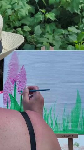 This media file captures an overhead, close-up shot of an individual painting outdoors. The subject, a person with visible shoulders and a portion of their back, is wearing a wide-brimmed straw hat and a black bra strap. They are holding a paintbrush in their right hand, actively applying strokes of pink paint to a canvas.

The canvas itself is propped on an easel and depicts a landscape scene. The background of the painting is a light gray with horizontal brushstrokes, suggesting a sky. In the foreground, green strokes represent grass, and several tall, elongated pink flower-like shapes, likely hyacinths, are already painted on the left side. The painter is adding details to these pink flowers.

The setting appears to be outdoors, amidst lush green foliage in the background, suggesting a garden or natural environment. The lighting indicates it's daytime, possibly morning or afternoon, with natural light illuminating the scene. There are no overt indications of weather, but the foliage suggests pleasant conditions.

A notable detail is the visible text "MALEVICH" on the wooden easel supporting the canvas. The overall mood is one of focused artistic creation in a serene, natural setting. The activity is clearly painting, with the artist engrossed in adding detail to the floral subject.