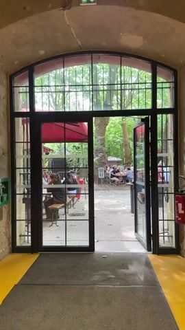 The camera slowly moves through a large black-framed doorway into an outdoor beer garden. The door is mostly glass, with a grid-like pattern of black bars over it, and it is open on the right side. Through the doorway, there are several people seated at picnic tables and round tables, chatting and enjoying drinks. A large red umbrella provides shade for one table. The area is surrounded by tall, leafy trees, some with colorful string lights hanging from them. The ground is a mix of concrete and gravel. The floor inside the doorway is concrete, with a grey rug and a yellow painted section.