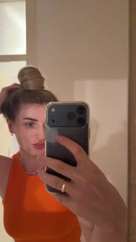 A woman with blonde hair styled in a bun is seen in a mirror taking a selfie. She is wearing an orange top and a gold ring on her left hand. Her hair is seen falling loose from the bun towards the end of the clip. She is holding a phone in her right hand, angled to capture her reflection. The background appears to be a room with a door and a light fixture visible.