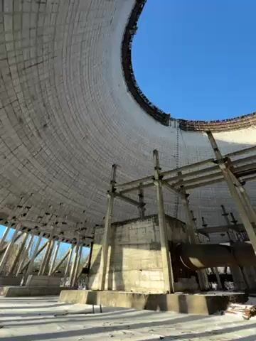 The video presents an interior view looking up inside a large, cylindrical, tower-like structure with a wide opening at the top, revealing a clear blue sky. The inner surface of the structure is lined with white or light-colored rectangular tiles, and there is a significant amount of deterioration and damage visible around the rim. A complex network of metal scaffolding and support structures are visible in the foreground and middle ground, rising towards the opening at the top. There are also large pipes and concrete blocks present, adding to the industrial look of the setting. The ground appears to be partially covered with snow.