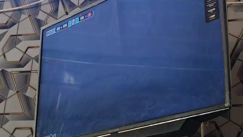 The video shows a television screen displaying a football match. The screen is angled slightly to the right, and the television is mounted on a wall covered with a patterned wallpaper. In the foreground, on the left side of the screen, a man wearing a black jersey with the number 10 and the Adidas logo is smiling. To his right, another man in a black shirt is also smiling and looking at the screen. In the center of the screen, a man wearing a blue and white striped jersey and a blue baseball cap is yelling enthusiastically. Behind him, a crowd of people in similar blue and white jerseys are cheering, with their arms raised. On the right side of the screen, a man in a black t-shirt with the Adidas logo is visible, also with his arm raised. The score visible on the screen shows 2-0. The overall atmosphere appears to be one of celebration and excitement among the football fans.