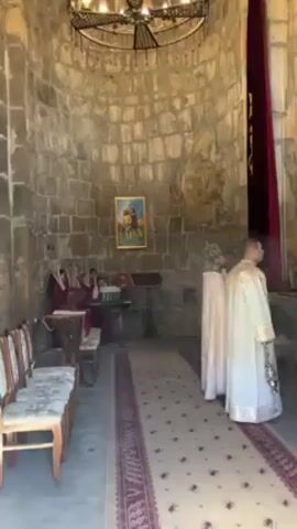 Inside an old stone church, a small choir sits to the left, dressed in maroon robes, accompanying their singing with an electronic keyboard. To the right, two men dressed in white and gold robes stand on a carpeted aisle, one holding a smoking censer, likely engaging in a religious ritual. The church has an aged stone dome, a chandelier hangs from the ceiling, and a painting adorns the wall above the choir. A maroon curtain hangs near the front of the church. The lighting is dim but natural, illuminating the interior.
