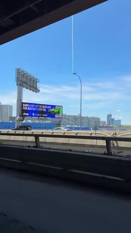 The video shows a highway from under a bridge. Cars drive on the highway and are visible through an opening between the bridge structure. A large billboard stands next to the highway. The billboard shows different advertisements, including a Jeep advertisement and one that says "CHECK IT, SWEAT IT". The sky is blue with some clouds and a contrail. Buildings are visible in the background. The bridge structure is made of steel and concrete.