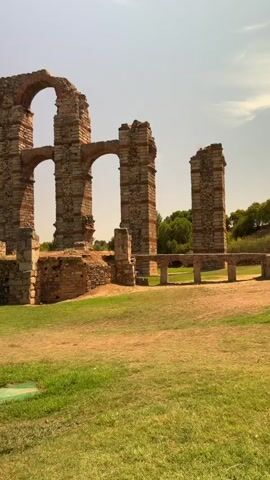 This is a vertical, medium shot video of the Acueducto de los Milagros, an ancient Roman aqueduct in Merida, Spain. The aqueduct is made of stone and brick, with large arches and pillars. The aqueduct is set against a backdrop of green grass and a blue sky with some clouds. The sun is shining brightly, casting shadows on the aqueduct. The video is filmed from a ground-level perspective, giving the viewer a sense of the scale and grandeur of the structure. There are no people or animals visible in the video.