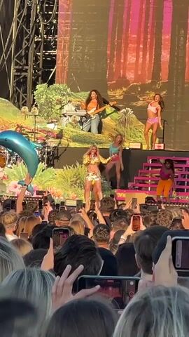 This is a vertical, slightly shaky video filmed from a crowd at a daytime outdoor concert. In the foreground, the heads and shoulders of many audience members are visible, most holding up smartphones to record or take pictures. In the mid-ground, on stage, there are several performers. The central performer, a blonde woman wearing a sparkly gold outfit, stands on a raised platform and is singing. To her right, another performer in a pink top and shorts dances on a staircase. Further back and higher up, two other women are performing, one playing a keyboard and the other dancing. A drum kit is visible to the left. A large inflatable blue dolphin is held up by someone in the crowd. The stage backdrop is a large screen displaying a colorful jungle or forest scene. The overall atmosphere appears energetic and festive.