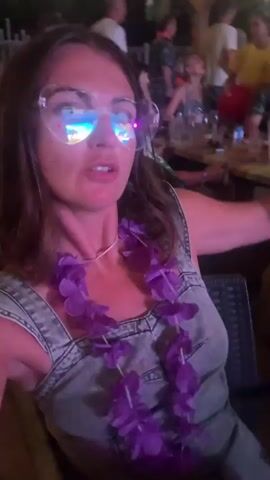A woman with brown hair, glasses, and a purple lei around her neck is filmed in what appears to be an outdoor restaurant or bar. She is wearing a denim overall dress. She is talking and making gestures with her hands. The background is blurred, but shows other people and tables, suggesting a social gathering. The lighting is dim and appears to be artificial, with purple hues visible on the woman.