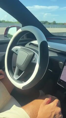 The video shows the interior of a Tesla car driving on a highway under a clear blue sky. A person's hands are visible on the steering wheel and in their lap. The steering wheel has a white cover with black textured grips. A touchscreen display on the dashboard shows navigation and driving information, including a graphic of the car and a semi-truck. The car is in the right lane, and other vehicles and an overhead highway sign indicating express lanes are visible in the distance.