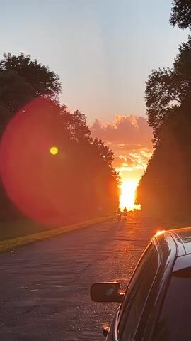 The video shows a sunset viewed from a car on a paved road. The sun is low on the horizon, casting a warm glow through the trees on either side of the road. Lens flare is visible in the upper left and right of the frame. Two people on motorcycles are riding away from the camera down the road. The car's side mirror and part of its roof are visible in the foreground.