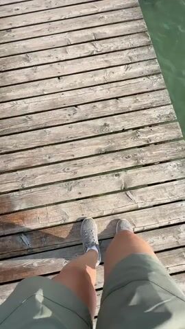 The video begins with a first-person perspective of someone walking on a wooden pier. They are wearing grey athletic shoes and green shorts. The pier is made of weathered wooden planks and extends over a body of green water. To the right of the pier, a metal ladder descends into the water, with some debris caught on it. The camera pans slightly to reveal more of the water and the surrounding landscape, which includes distant mountains and a cloudy blue sky. Another person, partially visible, is standing further down the pier. The video appears to be shot on a sunny day.
